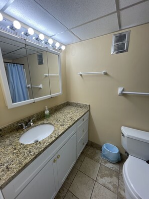 A bathroom with a granite countertop sink, a mirror with lights, a toilet, and a blue waste bin.