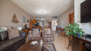 A spacious living room with a brown leather sofa, patterned armchairs, a wooden coffee table, and a dining area with wooden chairs and a table.