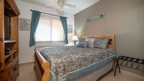 Une chambre à coucher comprenant un lit, une tête de lit en bois, un ventilateur de plafond, une fenêtre avec des rideaux, un panneau fixé au mur et une petite étagère avec quelques objets.