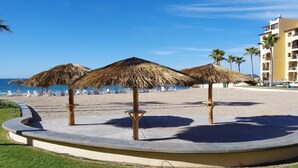 A beach with thatched umbrellas, white lounge chairs, and a building with balconies.
