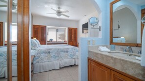 Une chambre à coucher avec un lit, un ventilateur de plafond, des volets en bois et une salle de bain équipée d’un lavabo et d’un miroir.
