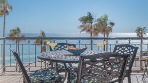 A round metal table with chairs, a bowl of fruit, and a drink on a balcony overlooking the ocean.