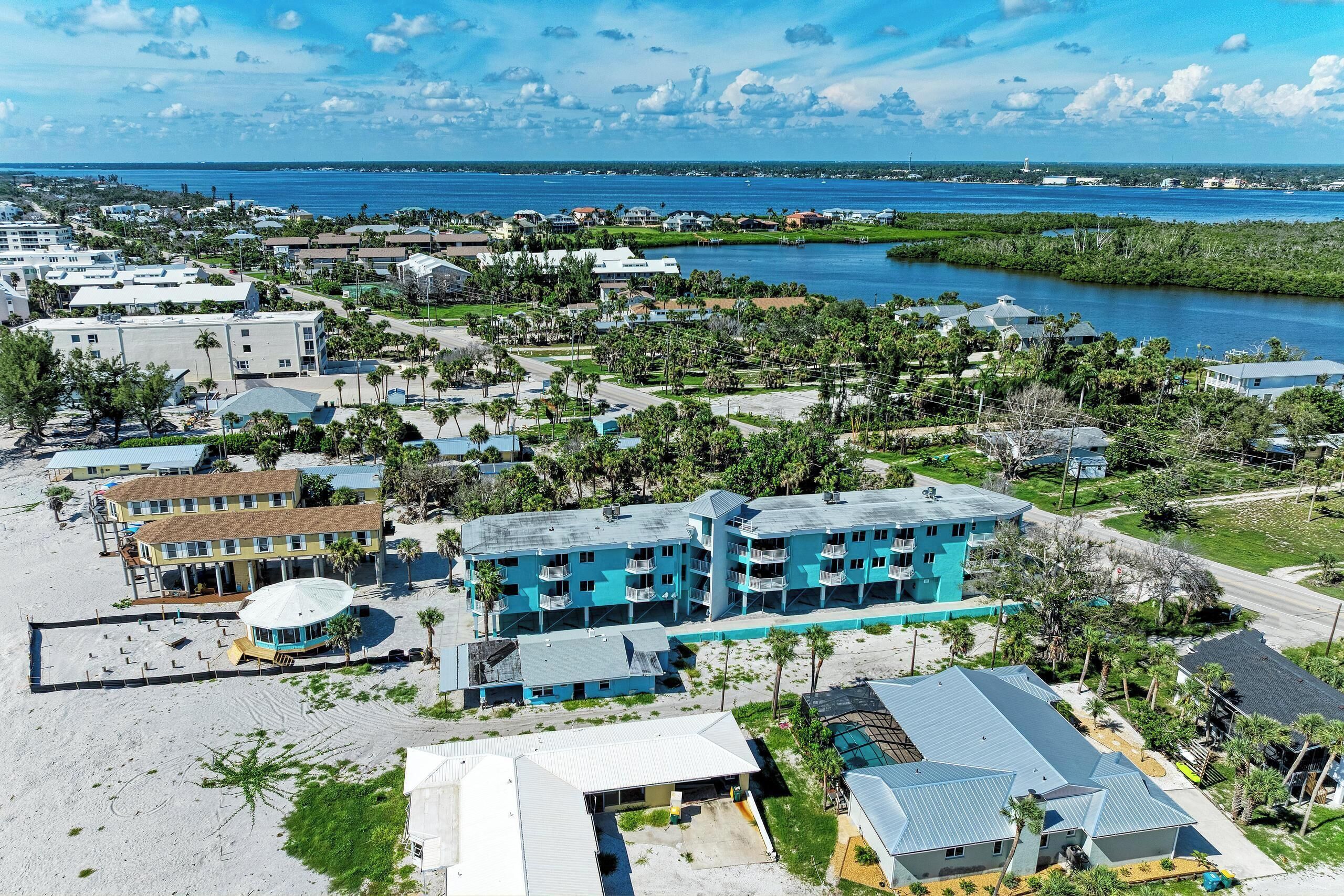 An aerial view of a coastal residential area with multiple buildings, including a prominent blue building with balconies, surrounded by greenery and a body of water.