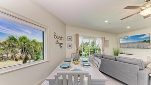 Un coin repas avec vue sur les palmiers et la plage, un canapé gris et un ventilateur de plafond.