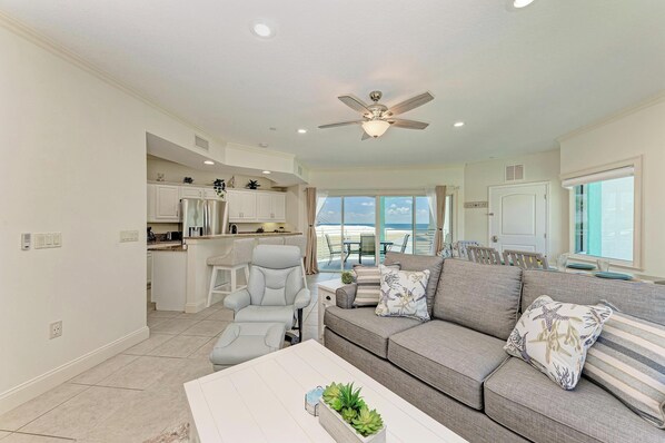 A spacious living room with a gray sofa, a white coffee table, and a ceiling fan. A kitchen with white cabinets and a dining area with a table and chairs is visible in the background.