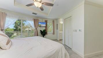 A bedroom with a large bed, a ceiling fan, a TV, and a view of the outdoors.
