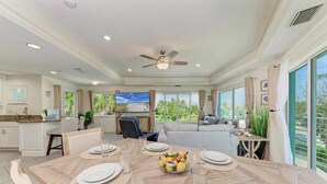 Une salle à manger spacieuse avec une grande table dressée pour le repas, un ventilateur de plafond et une cuisine en arrière-plan.