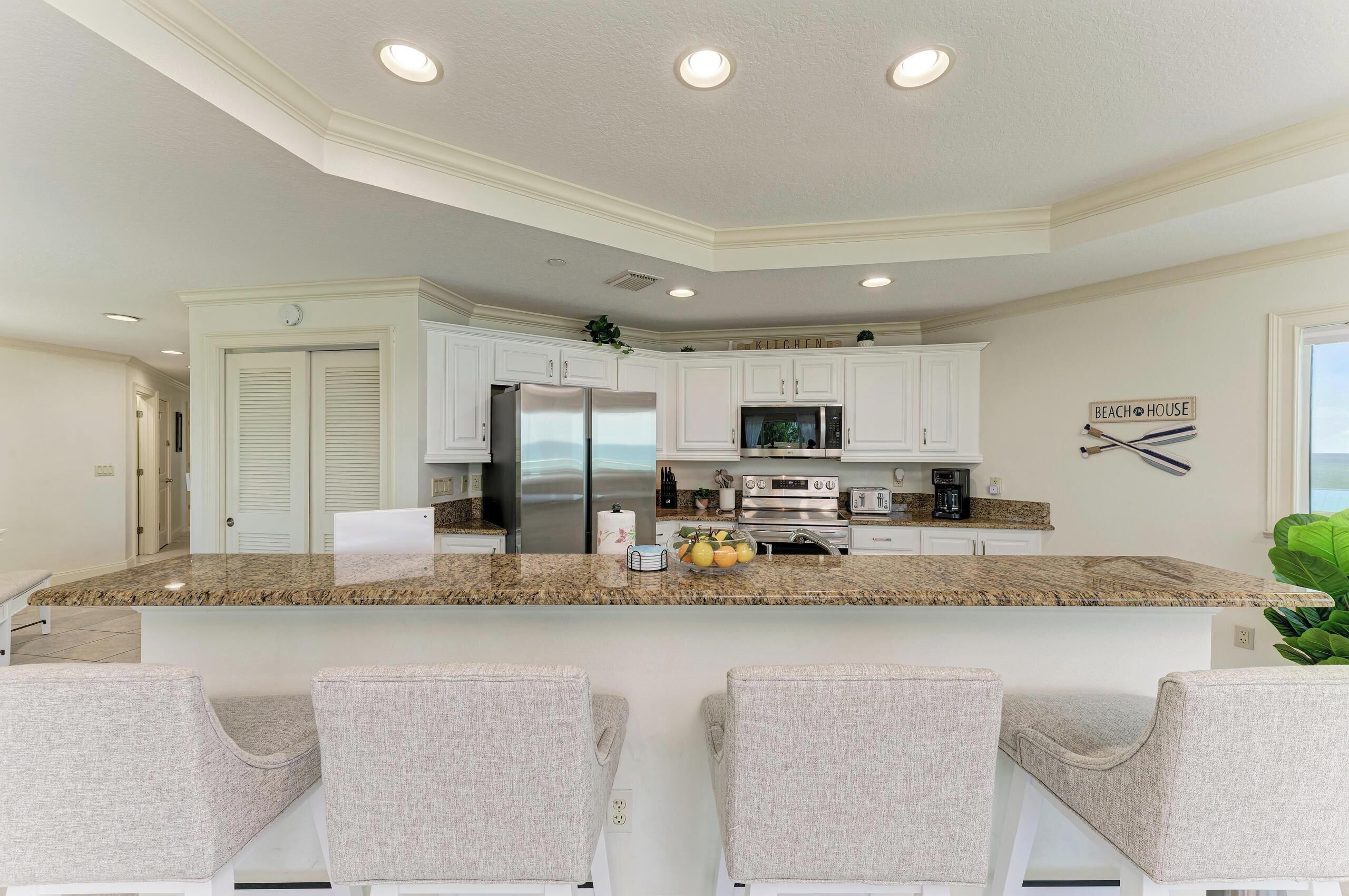 Une cuisine moderne avec des meubles blancs, un plan de travail en granit, des appareils électroménagers en acier inoxydable et un coin bar doté de chaises rembourrées.