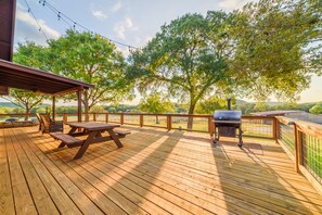 Una terraza de madera con mesas de picnic y una parrilla, rodeada de árboles y vistas al paisaje.