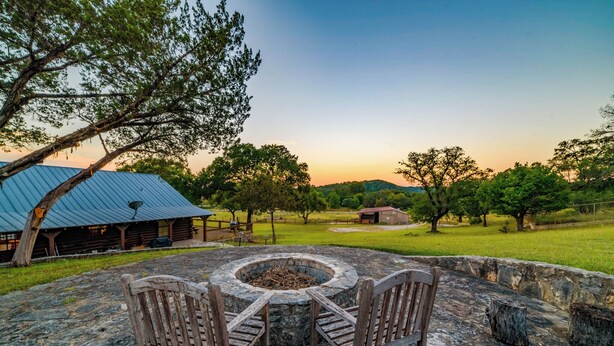A stone fire pit with two wooden chairs, surrounded by a stone wall, with a view of a rustic building and a grassy field in the background. - River l Resort Pool l Lake l Firepit l HOT TUB (Ingram)