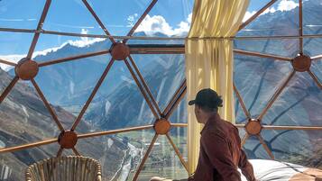 A room with a bed, a chair, and a view of mountains through a geometric window structure.
