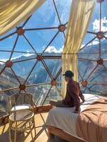 Una habitación con una cama, una silla y vistas a montañas a través de una estructura de ventana geométrica.