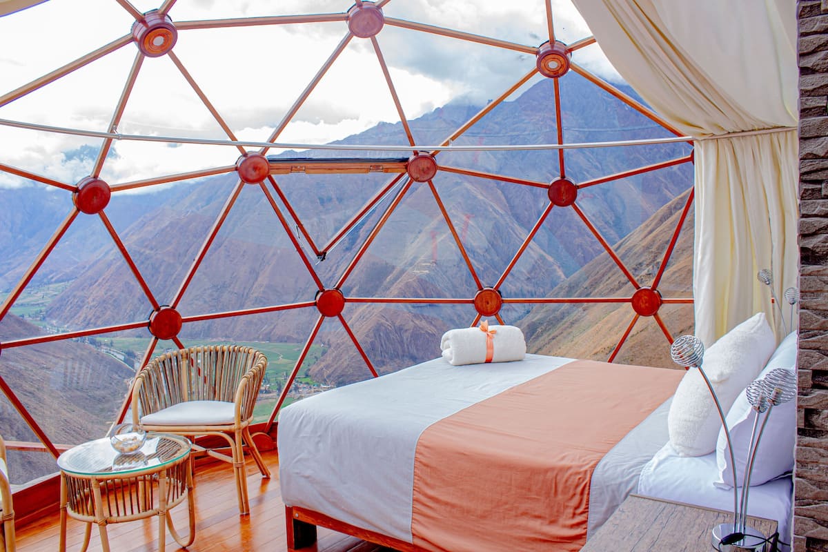 A room with a bed, a chair, and a table, with a mountain view through a geometric window structure.