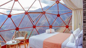 Habitación con cama, silla y mesa, con vista a la montaña a través de una estructura de ventana geométrica.