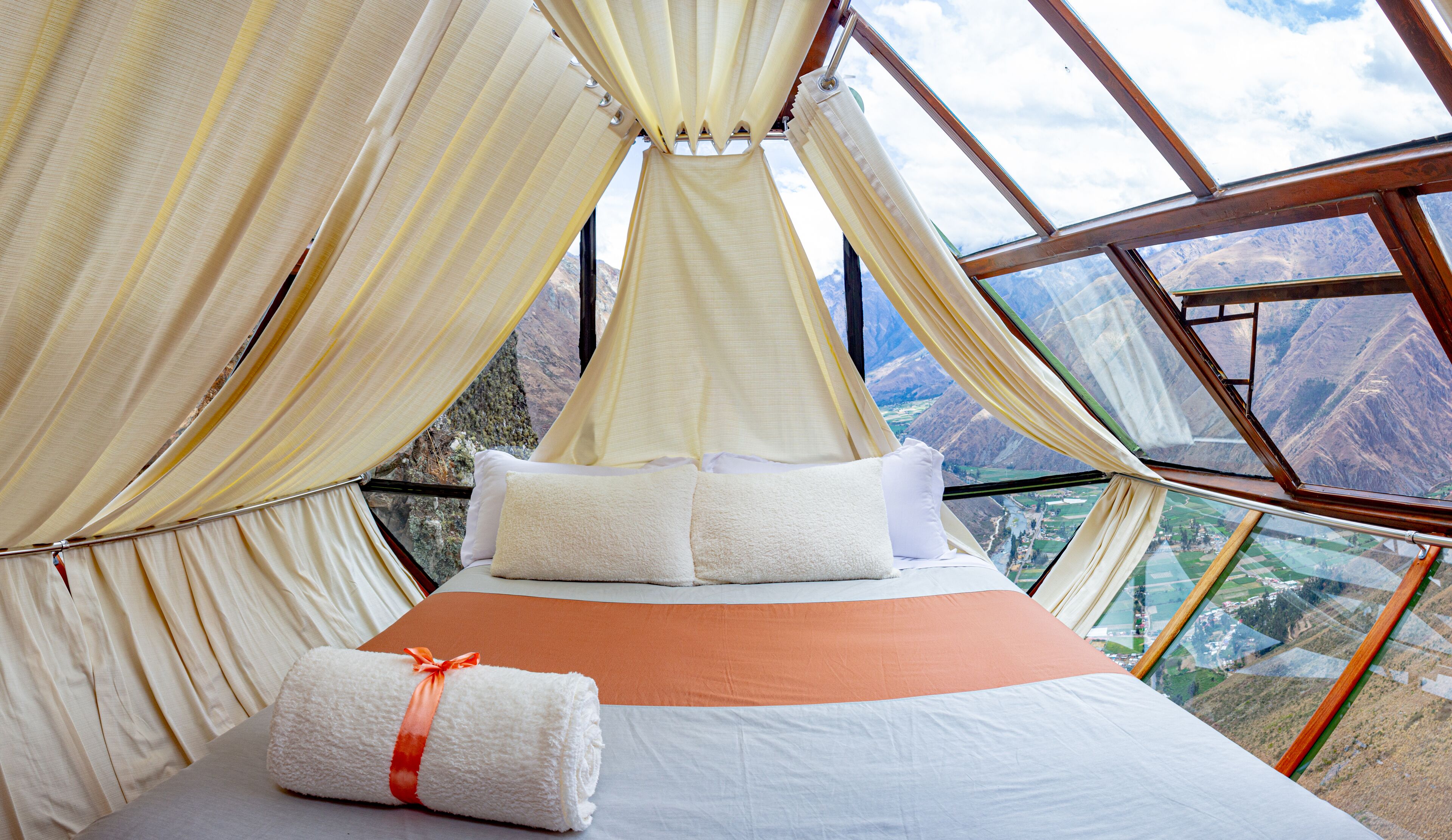 Una cama con sábanas y almohadas blancas, una manta marrón y una toalla enrollada con un lazo naranja, con vistas a montañas a través de un amplio ventanal.
