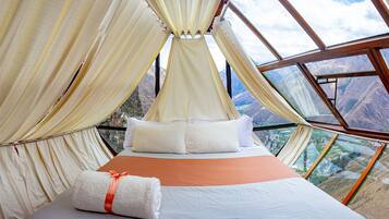 Una cama con sábanas y almohadas blancas, una manta marrón y una toalla enrollada con un lazo naranja, con vistas a montañas a través de un amplio ventanal.