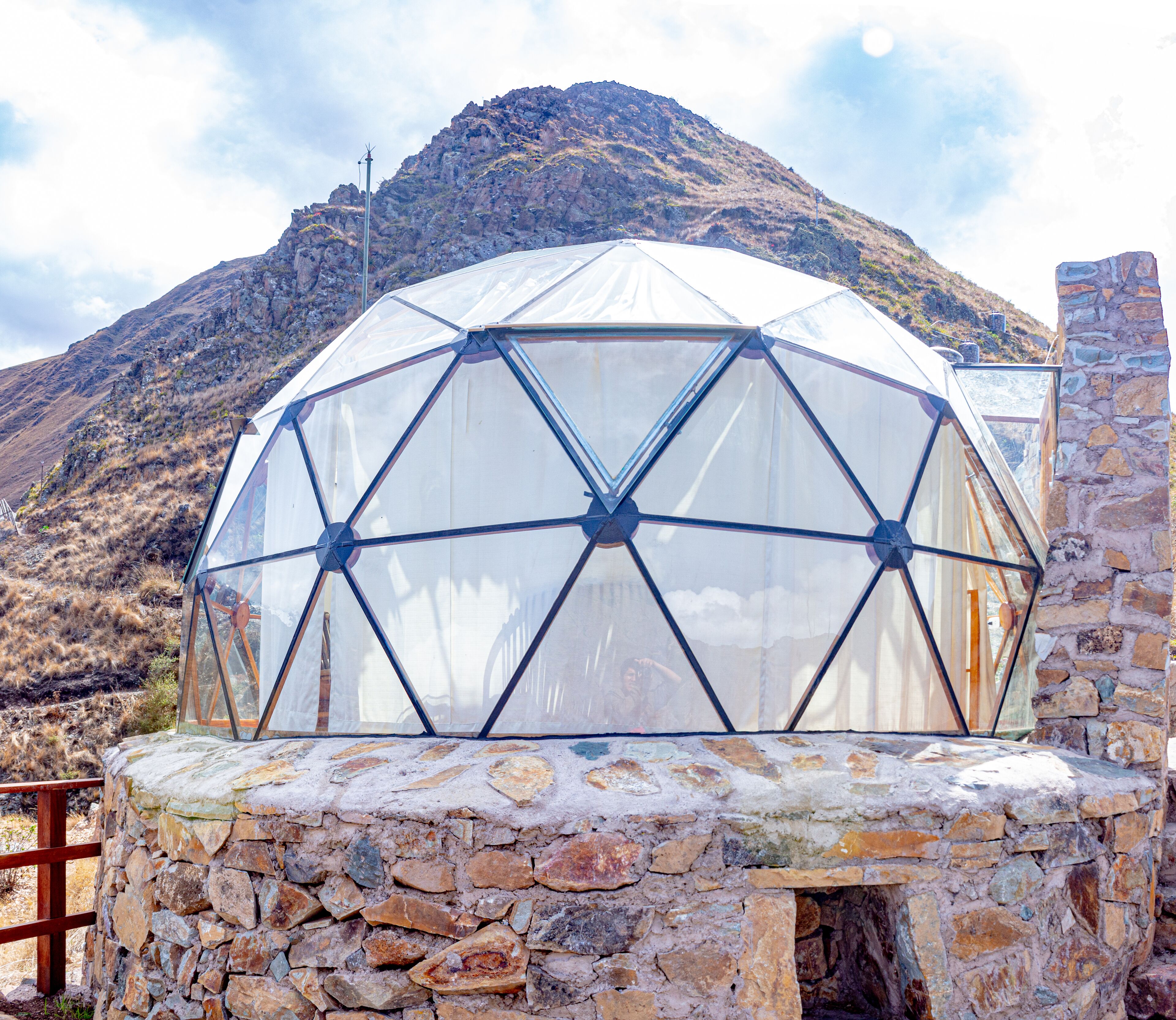 A geodesic dome structure with a stone base, set against a mountainous backdrop.