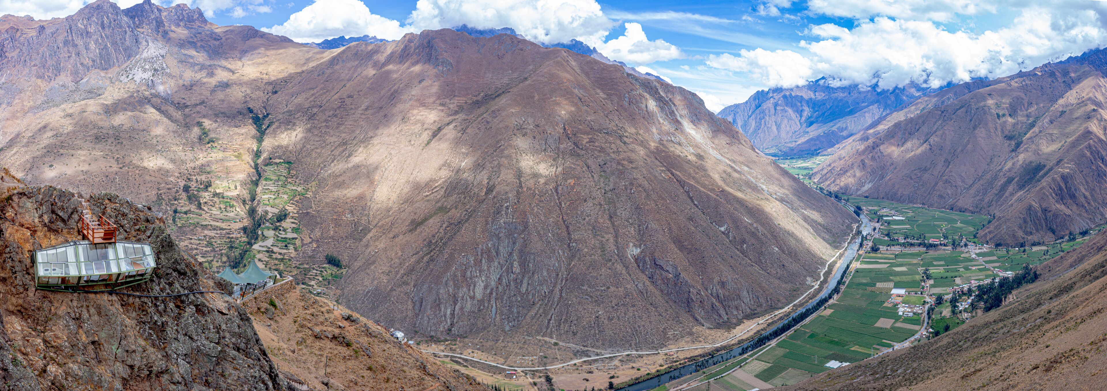 Un paisaje montañoso con un valle, un río y una estructura tipo invernadero sobre un acantilado.