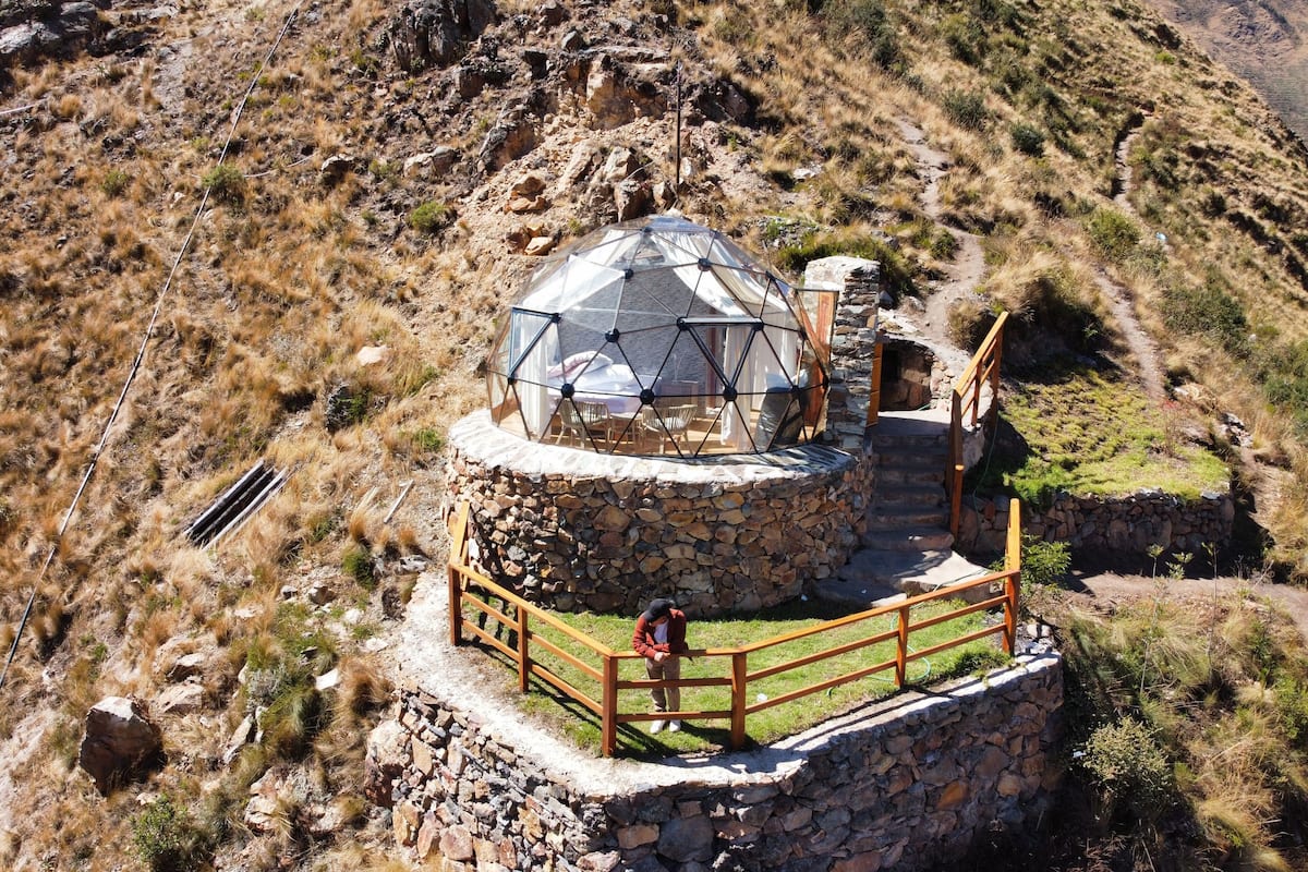 A geodesic dome structure with a stone base, surrounded by a wooden fence, set on a hillside.