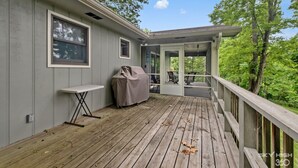 A covered outdoor deck with a table and chairs, a barbecue grill, and a view of the surrounding trees.