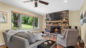 A living room with a stone fireplace, a ceiling fan, and a large window overlooking a green landscape.