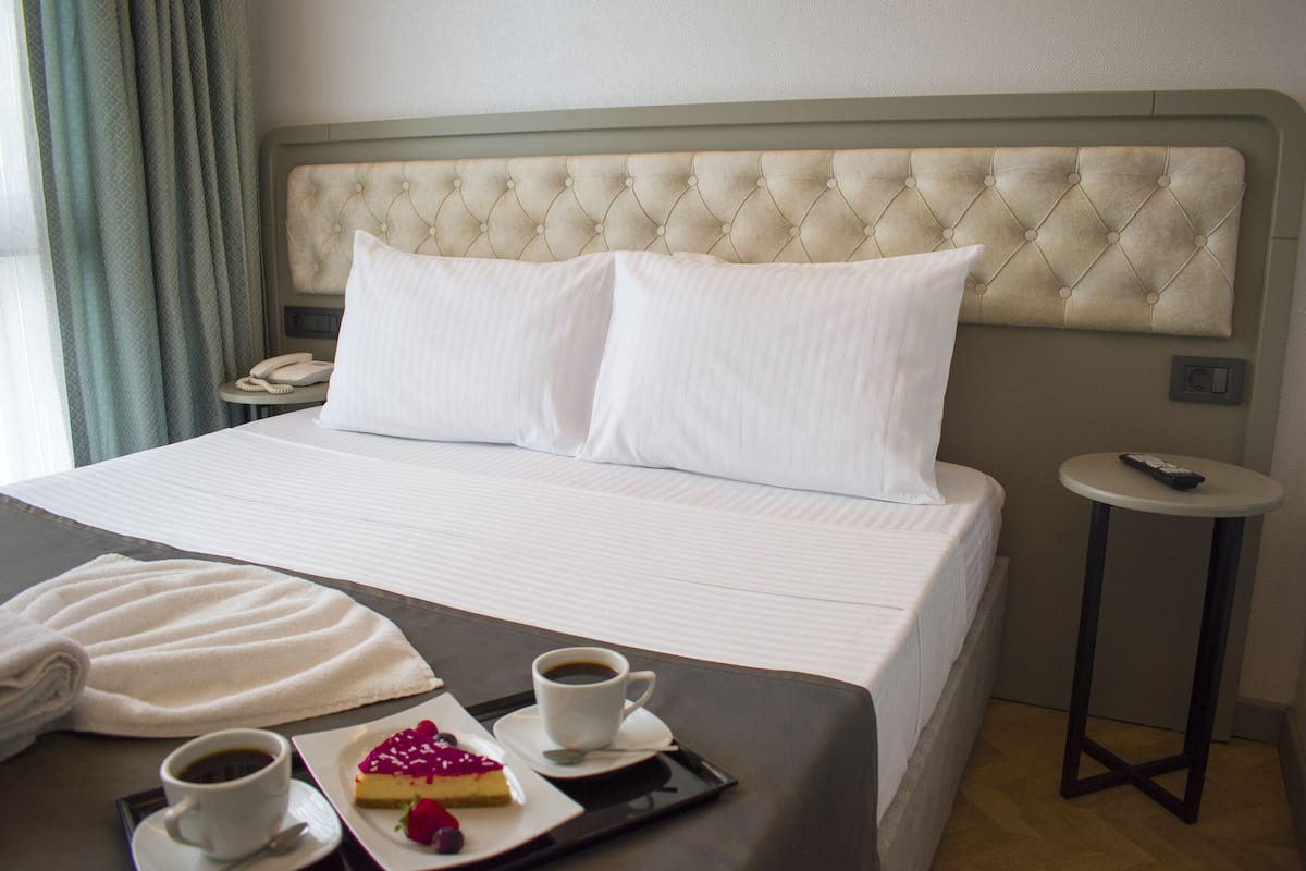 Ein ordentlich bezogenes Bett mit weißen Leinentüchern, ein Nachttisch mit einem Telefon und ein Tablett mit einem Kuchenstück und zwei Tassen Kaffee.
