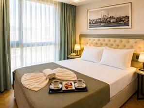 Une chambre d’hôtel avec un grand lit, un tableau représentant un paysage urbain, une fenêtre avec des rideaux légers et un plateau avec un dessert et du café.