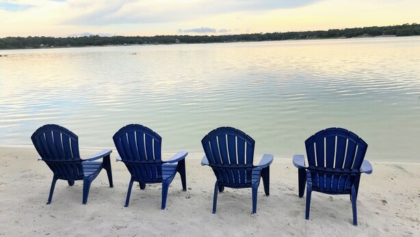 Tres cadires Adirondack blaves davant d'un llac tranquil amb una riba llunyana.