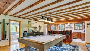 Una mesa de billar en una habitación con vigas de madera, una zona de cocina y un comedor con sillas.