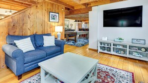 Una sala de estar acogedora con un sofá azul, una mesa de centro blanca, vigas de madera, un televisor y un estante con diversos objetos.