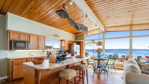 A kitchen with wooden cabinets, a large island with stools, and a view of the ocean through large windows. - Wonderful Waterfront Escape w/ an Indoor Hot Tub, Furnished Deck, & Great Views (Clinton)
