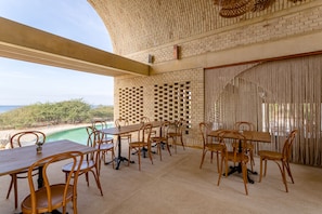 A dining area with a long table and chairs, a brick wall, and a view of the beach.