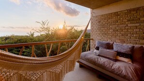 A hammock on a balcony with a view of the sunset over the ocean, a brick wall, and a sofa with cushions.
