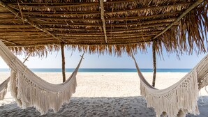 A hammock under a thatched roof on a sandy beach with the ocean in the background.