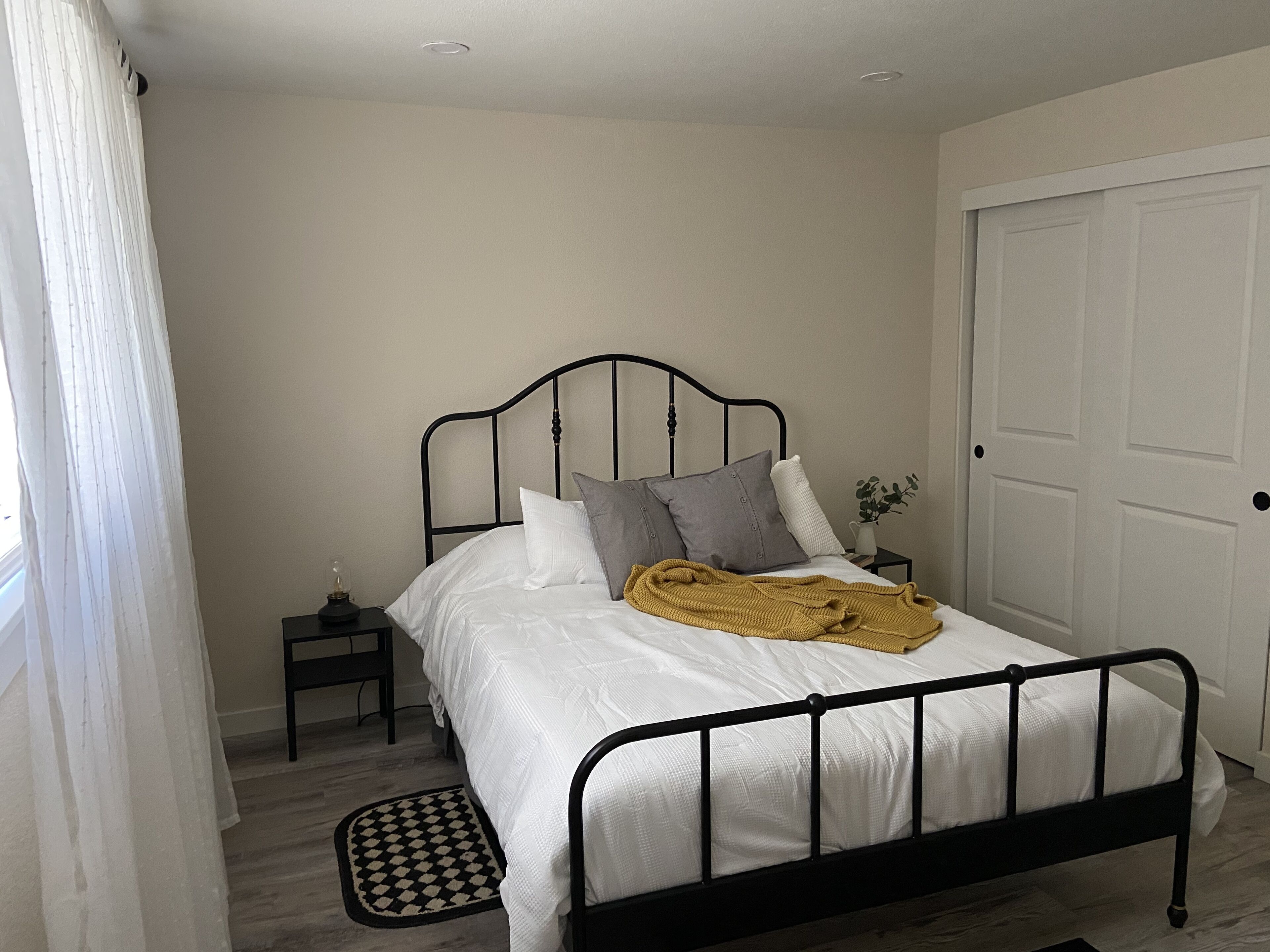 A neatly made bed with white bedding and a mustard-colored throw blanket, a small bedside table with a lamp, and a door to the right.