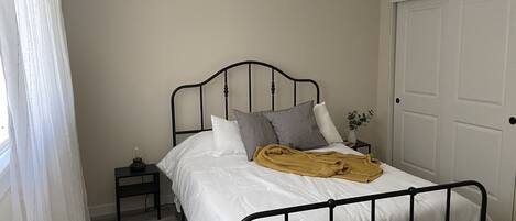 A neatly made bed with white bedding and a mustard-colored throw blanket, a small bedside table with a lamp, and a door to the right.
