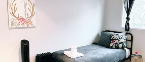 A small, modern bedroom with a single bed, a radiator, a window with curtains, and a wall-mounted artwork.
