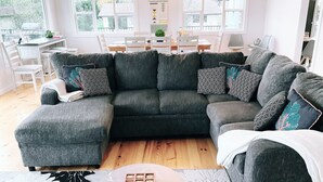 Ein modernes Wohnzimmer mit einem Ecksofa, einem Esstisch und Stühlen. Eine Küchenzeile ist durch das Fenster erkennbar, inklusive eines Grills.