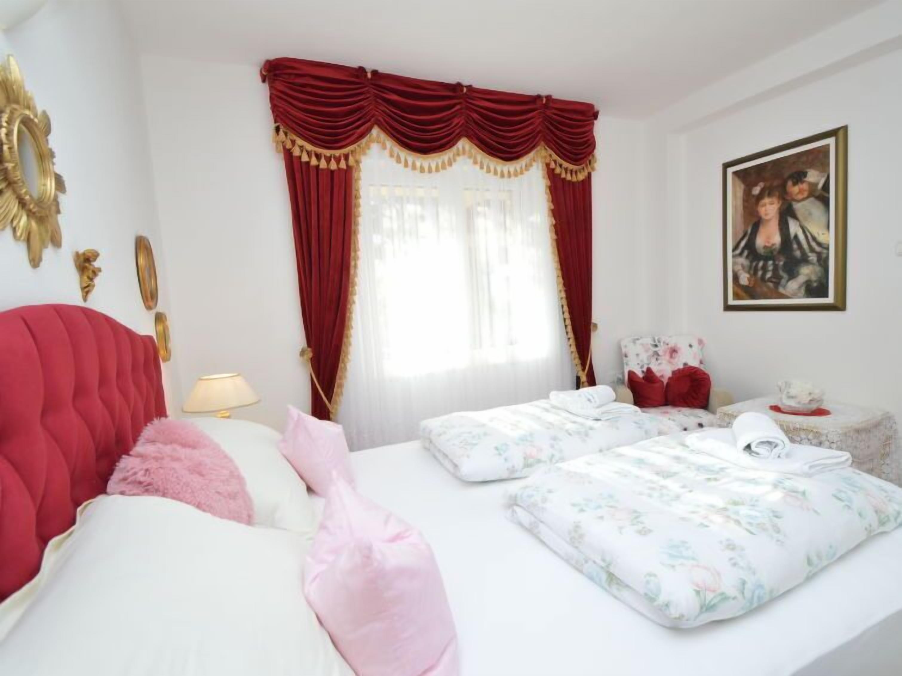 A hotel room with a red upholstered headboard, a bed with floral bedding, a bedside table with a lamp, and a painting on the wall.