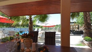 An outdoor patio with wooden tables, chairs, and umbrellas. There are palm trees and a pool in the background.
