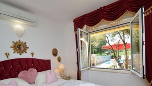 A bedroom with a large bed, a window with red curtains, and a view of an outdoor swimming pool.