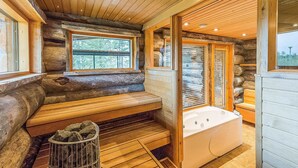 A log cabin interior with a stone fireplace, a wooden bench, a bathtub, and a window with a view of trees. - Jussis chalet a by Interhome (Kuusamo)