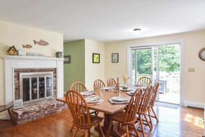 A dining room with a brick fireplace, wooden chairs, a dining table set for six, and a view through a glass door to the outdoors.