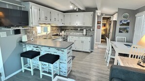 Een moderne keuken met witte kasten, een centraal eiland met barkrukken, een eethoek met een tafel en stoelen, en een televisie boven het eiland.