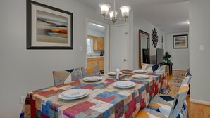 Een eetruimte met een lange tafel voor zes personen, een kroonluchter en een lijst met een abstract schilderij aan de muur.