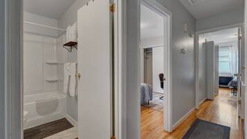 A modern bathroom with a shower, toilet, and towel rack. A hallway leads to a bedroom with a bed and a chair.