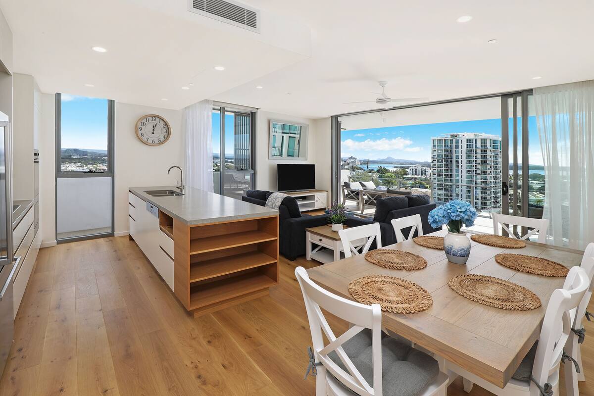 Una cocina moderna con una isla grande, un comedor con mesa de madera y sillas blancas, y un ventanal con vista a la ciudad.