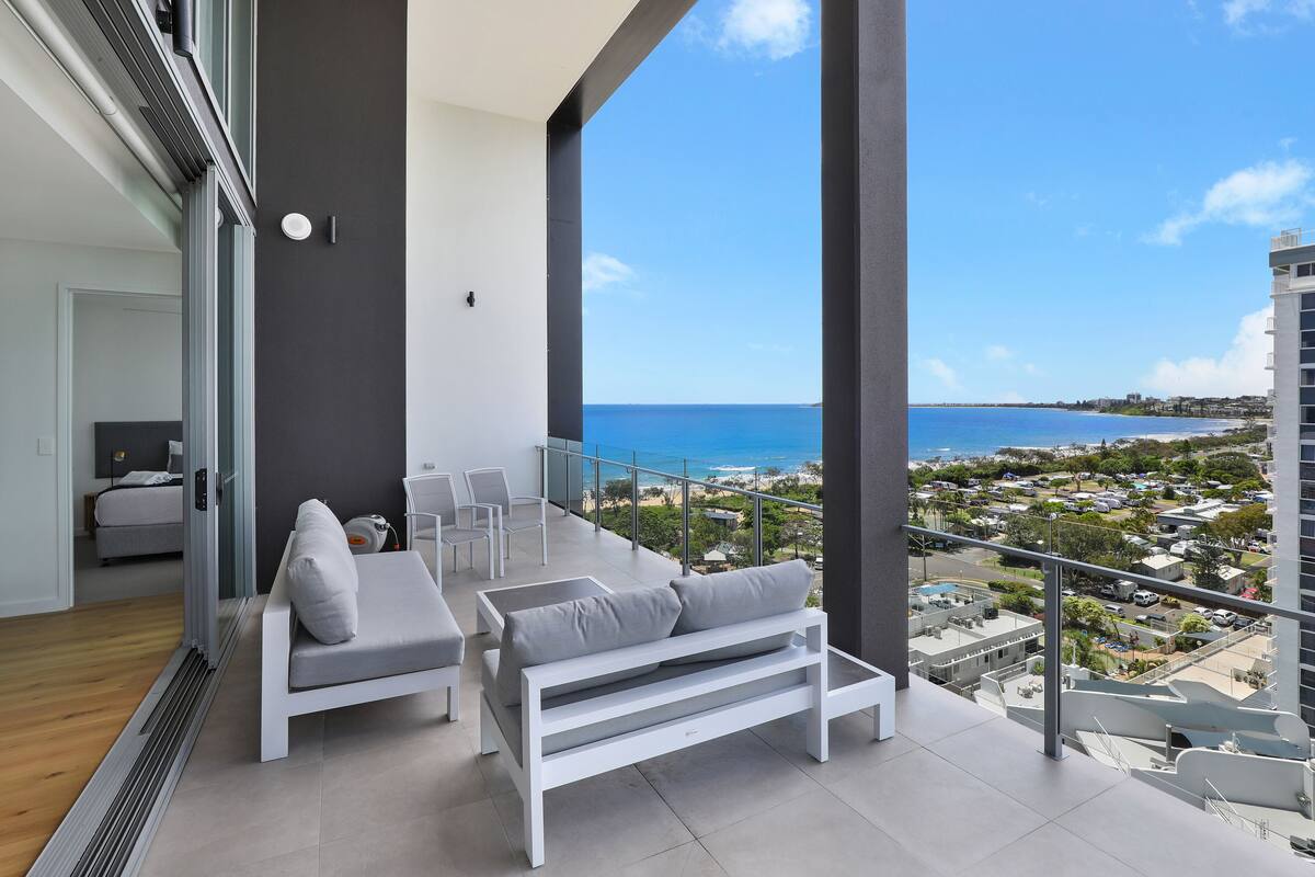 Un balcón con sofá y sillones blancos, vistas al mar y un paisaje urbano de fondo.