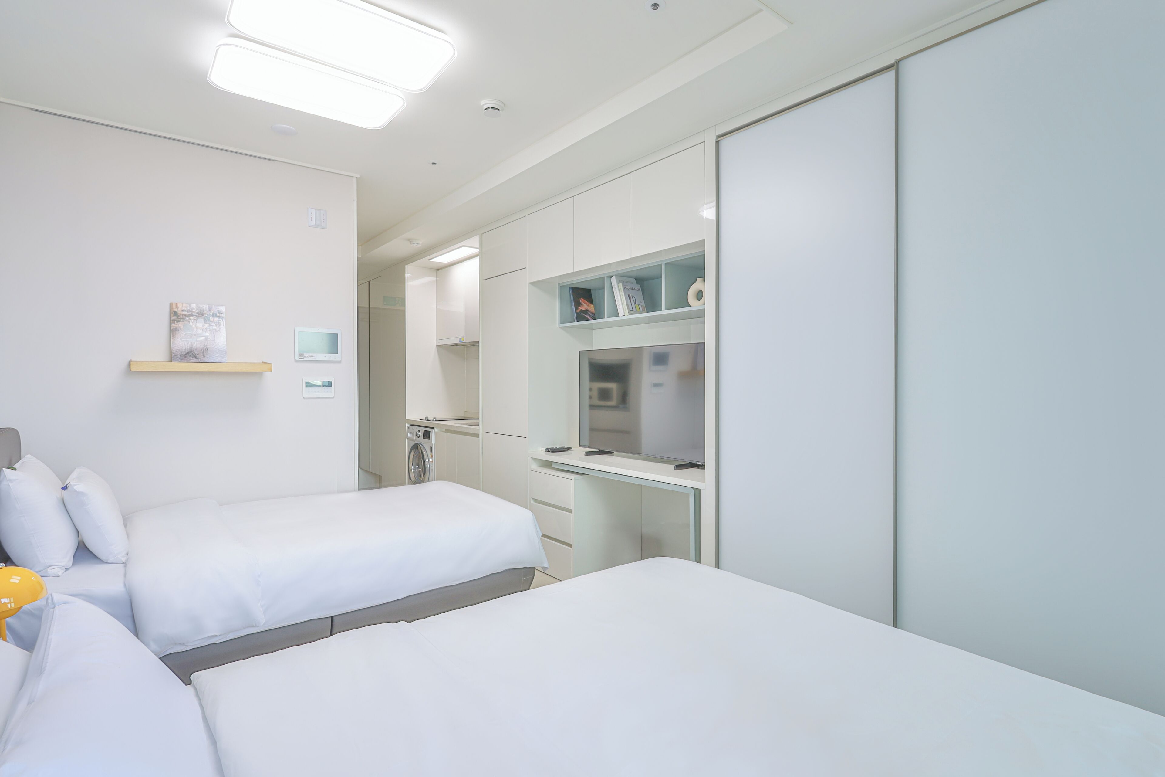 A modern hotel room with two beds, a desk, a mirror, and a small kitchenette.