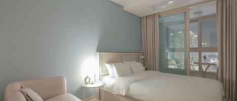 A hotel room with a large bed, a small side table, a sofa, and a view of a cityscape through large windows.